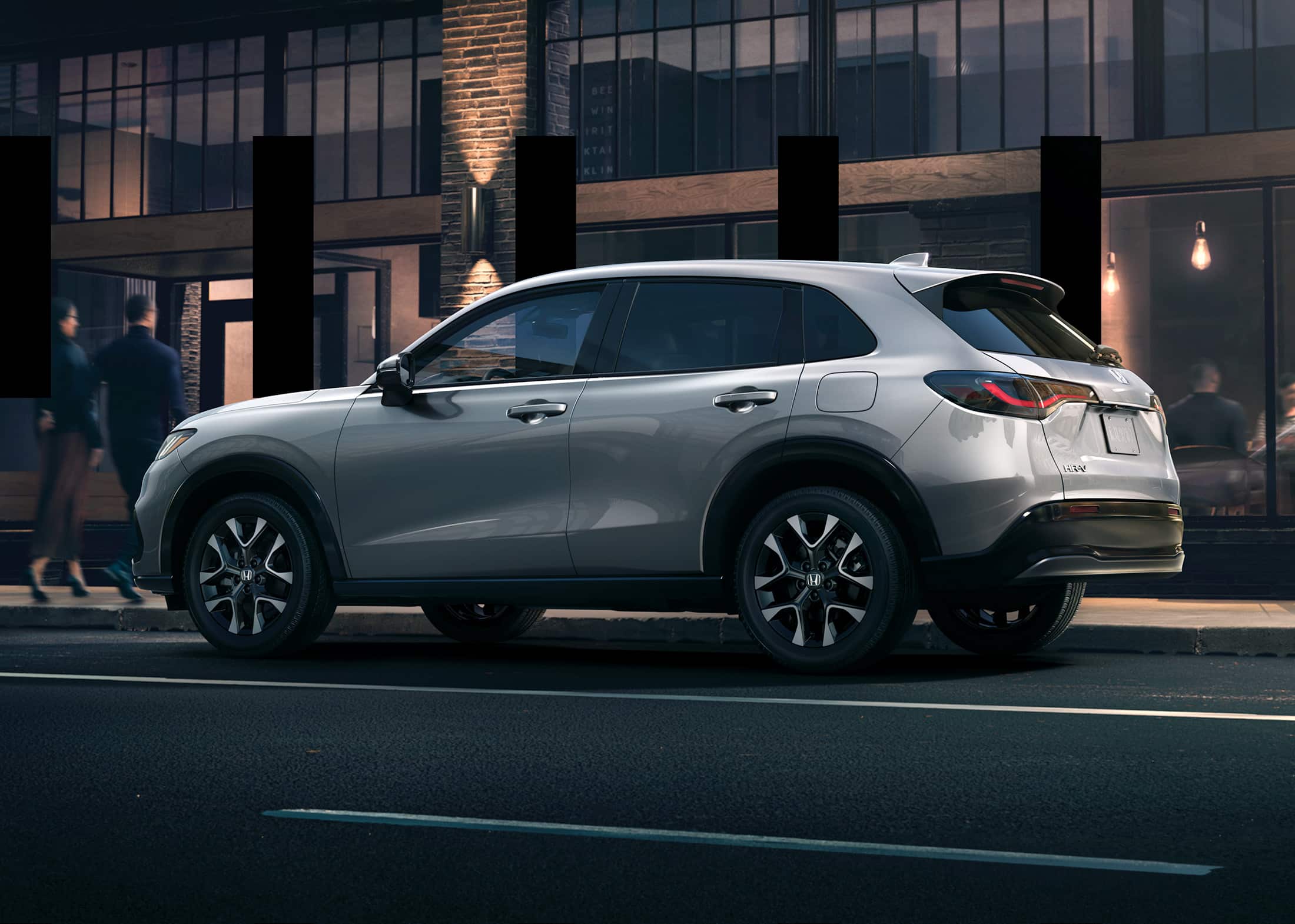 7/8 rear driver-side view of the 2026 Honda HR-V EX-L in Solar Silver Metallic, shown parked in front of a restaurant at night. 