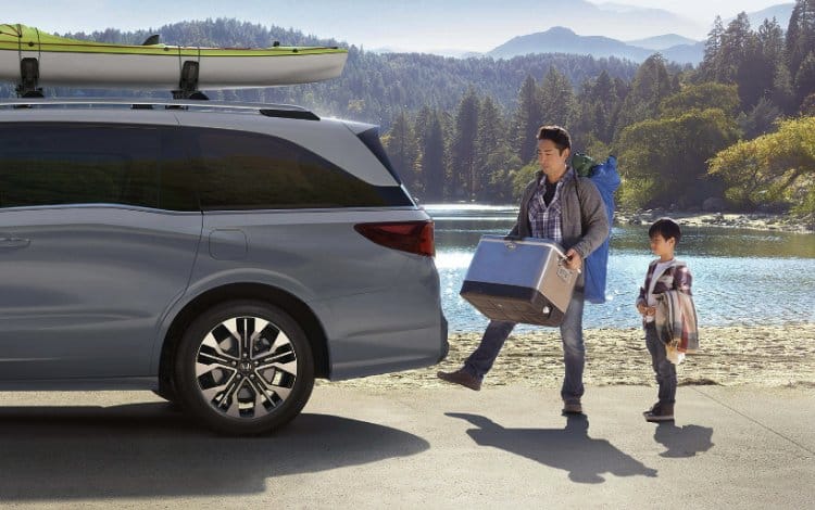 2026 Honda Odyssey Elite parked at lake with hills and greenery in background. Father using the motion sensor to open the back door to load items while son is watching.